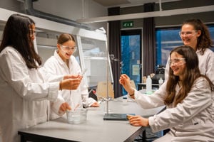 4 jenter i kvite frakkar sit rundt eit bord og gjera eit kjemisk forsøk