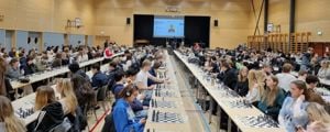 Ein gymsal full av elevar som sit ved langbord og speler simultansjakk
